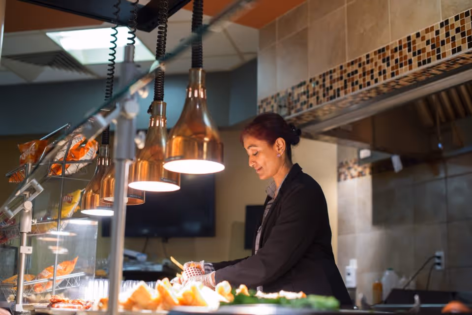 A staff member stands behind a buffet serving counter under copper heat lamps, preparing and arranging food.