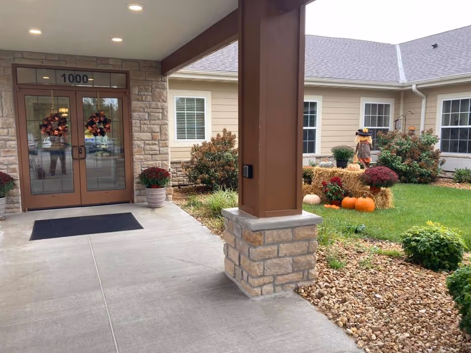 Covered building entrance with glass double doors labeled "1000", stone pillars, and fall decorations including pumpkins and hay bales on the front lawn.