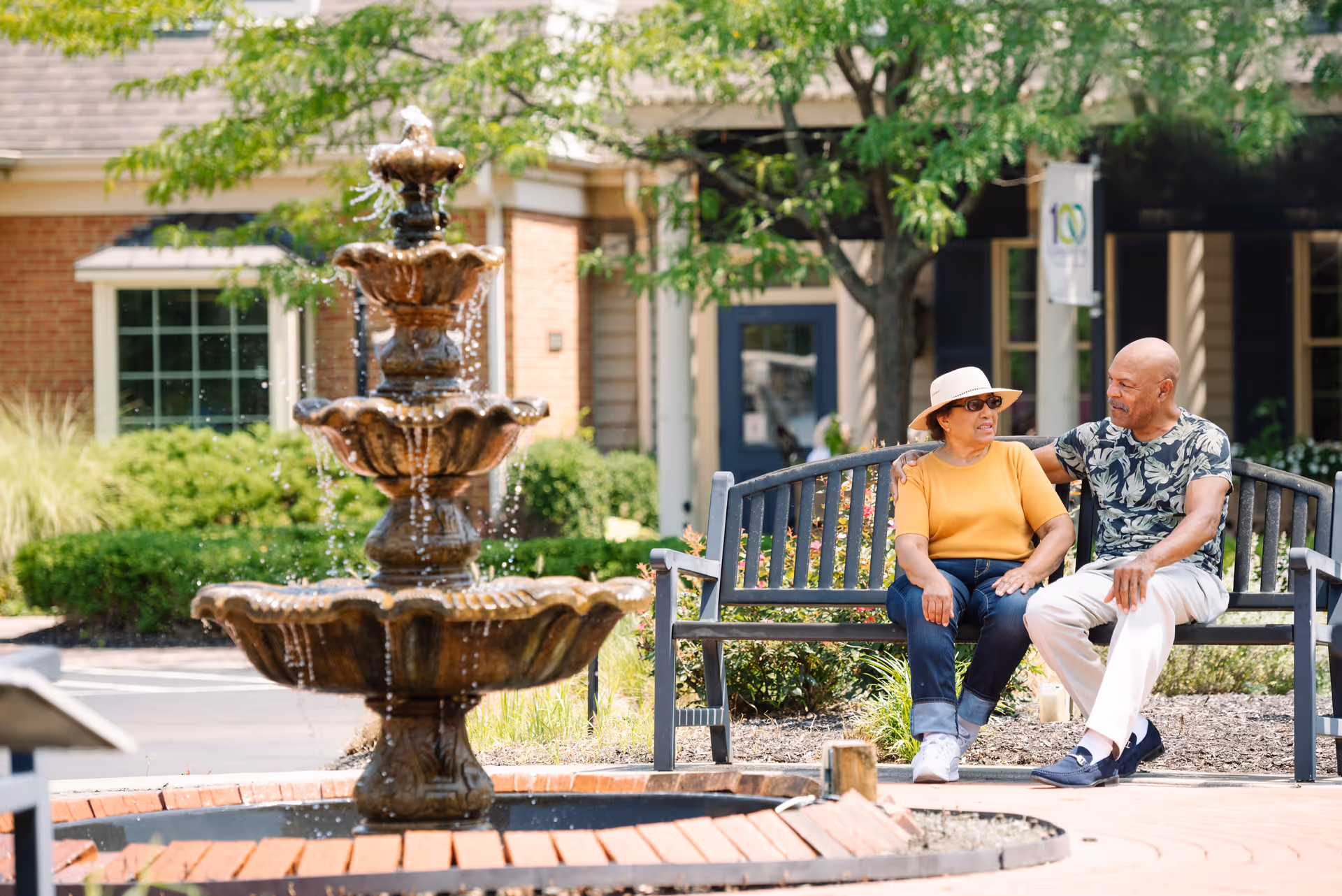 An elderly couple sitting on a bench near a multi-tiered water fountain in a garden area outside a residential building. The woman is wearing a hat, sunglasses, a yellow shirt, and jeans, while the man is wearing a patterned shirt and light-colored pants. They appear to be engaged in conversation on a sunny day.