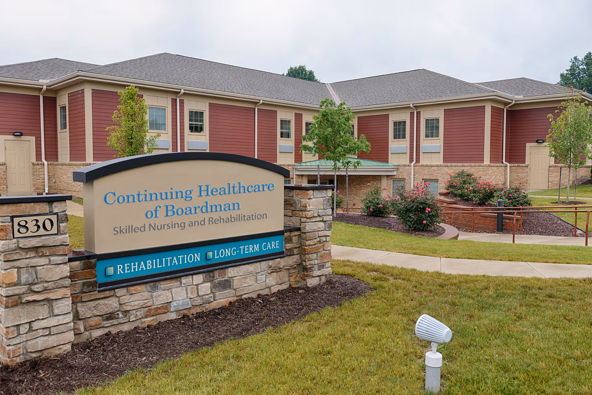 Exterior view of Continuing Healthcare of Boardman facility showing a stone and brick sign with the facility name and services offered, surrounded by grass, shrubs, and a paved walkway leading to the building entrance.