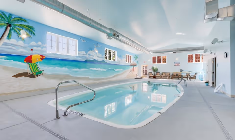 Indoor swimming pool area with a beach-themed mural on the wall depicting a sandy shore, ocean waves, palm trees, and a colorful beach umbrella with a lounge chair. The pool has metal handrails and a 'No Diving' warning on the edge. There are several windows letting in natural light and a seating area with chairs and plants at the far end of the room.