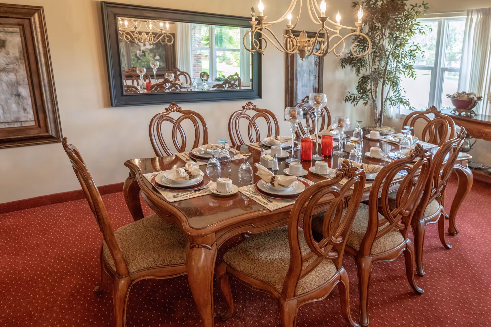 Elegant dining room with a set wooden table, carved chairs, chandelier, and place settings on a red carpeted floor.