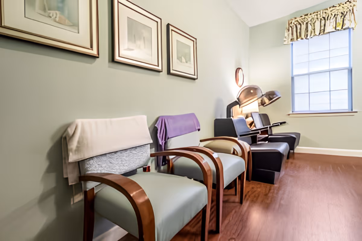 A room with several chairs lined up against a light green wall, two of which have towels draped over the backs. There are three framed pictures hanging on the wall above the chairs. In the background, there are two black salon-style chairs with hair dryers attached, positioned near a window with a valance curtain. The floor is wooden.
