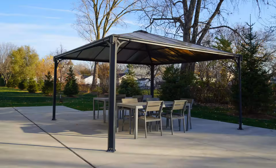 Metal gazebo over an outdoor dining table and chairs on a concrete patio with trees and lawn in the background.