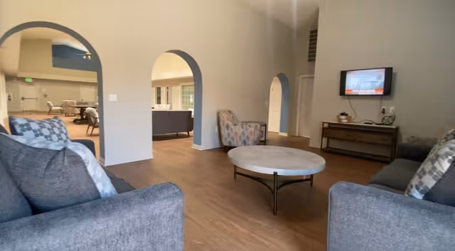 Communal living room with sofas, armchairs, a round coffee table, wall-mounted TV, and arched doorways.
