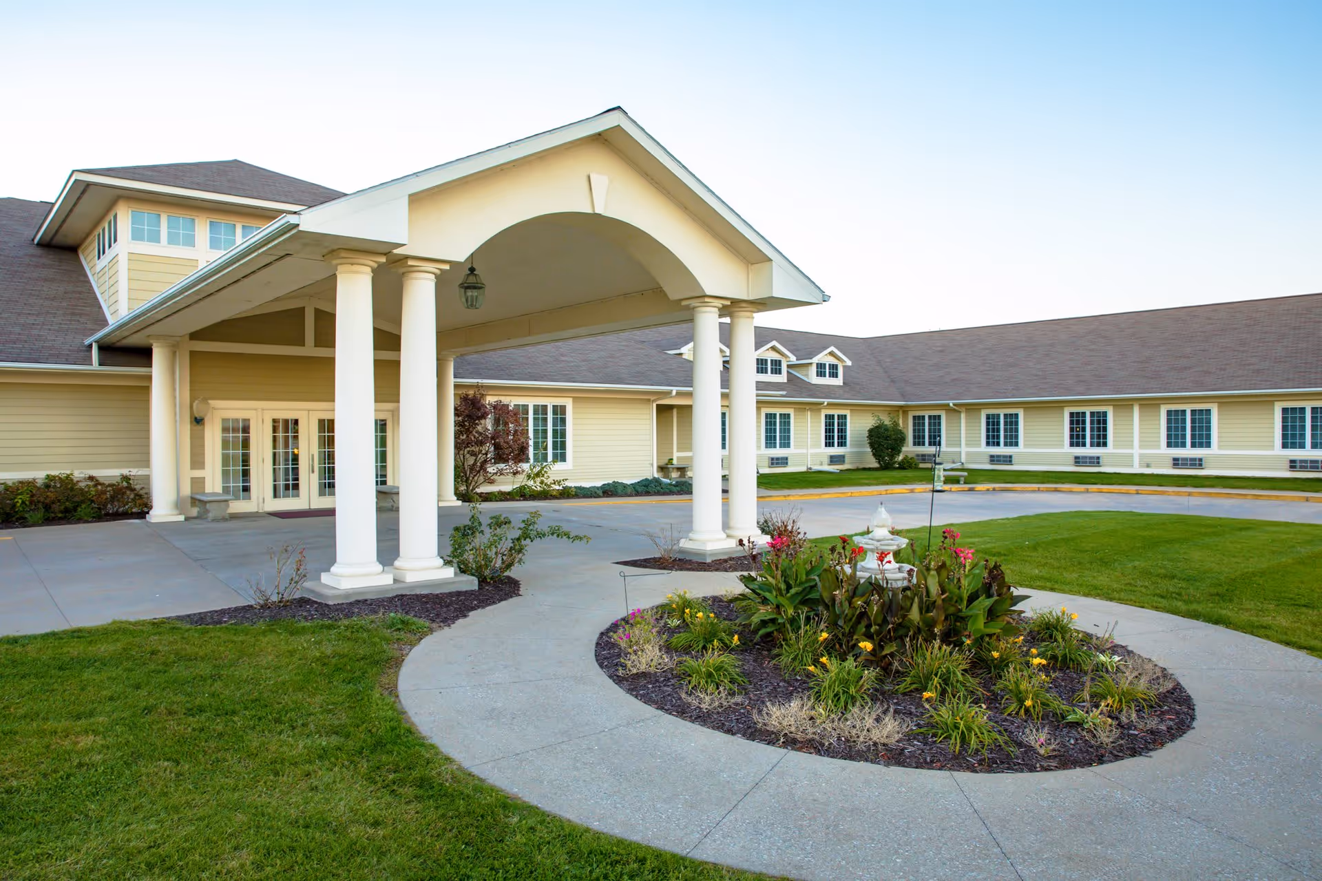 Exterior view of a single-story assisted living facility with a covered entrance supported by white columns. The building is light yellow with white trim and has multiple windows. In front of the entrance is a circular driveway with a landscaped garden featuring various plants and flowers.