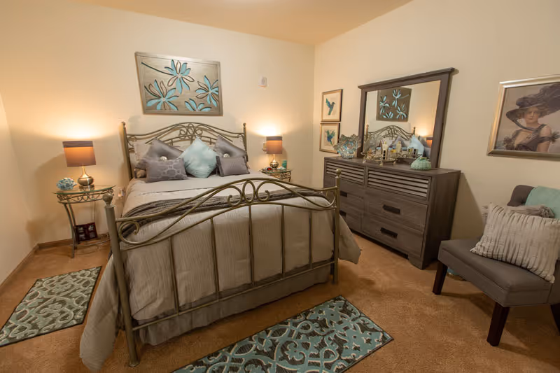 A cozy bedroom with a metal frame bed dressed in beige and light blue bedding, two matching bedside tables with lamps, a wooden dresser with a mirror, decorative artwork on the walls, a cushioned chair with pillows, and patterned rugs on a carpeted floor.