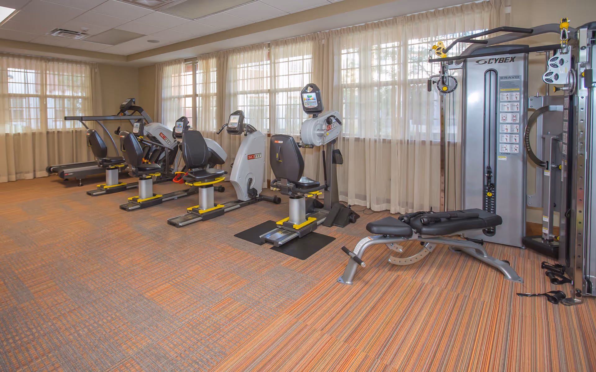 A fitness room with exercise equipment including treadmills, recumbent bikes, a rowing machine, and a multi-function weight machine. The room has large windows with sheer curtains allowing natural light to fill the space, and the floor is covered with patterned carpet tiles.