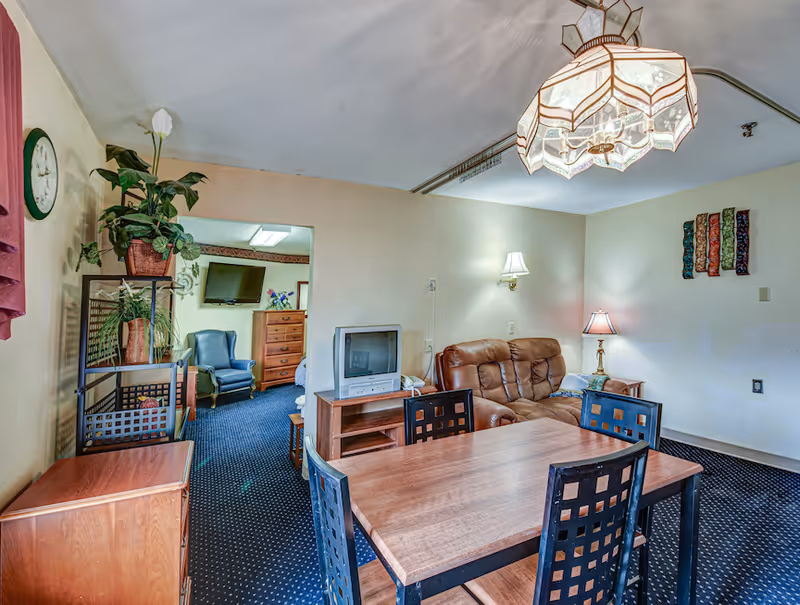 Interior view of a senior living facility room with a wooden dining table and four chairs in the foreground. Behind the table is a brown leather couch, a small wooden TV stand with an older model television, and a side table with a lamp. To the left, there is a shelving unit with potted plants and a wall clock. In the background, another room is visible with a blue armchair, a wooden dresser, and a wall-mounted flat screen TV.