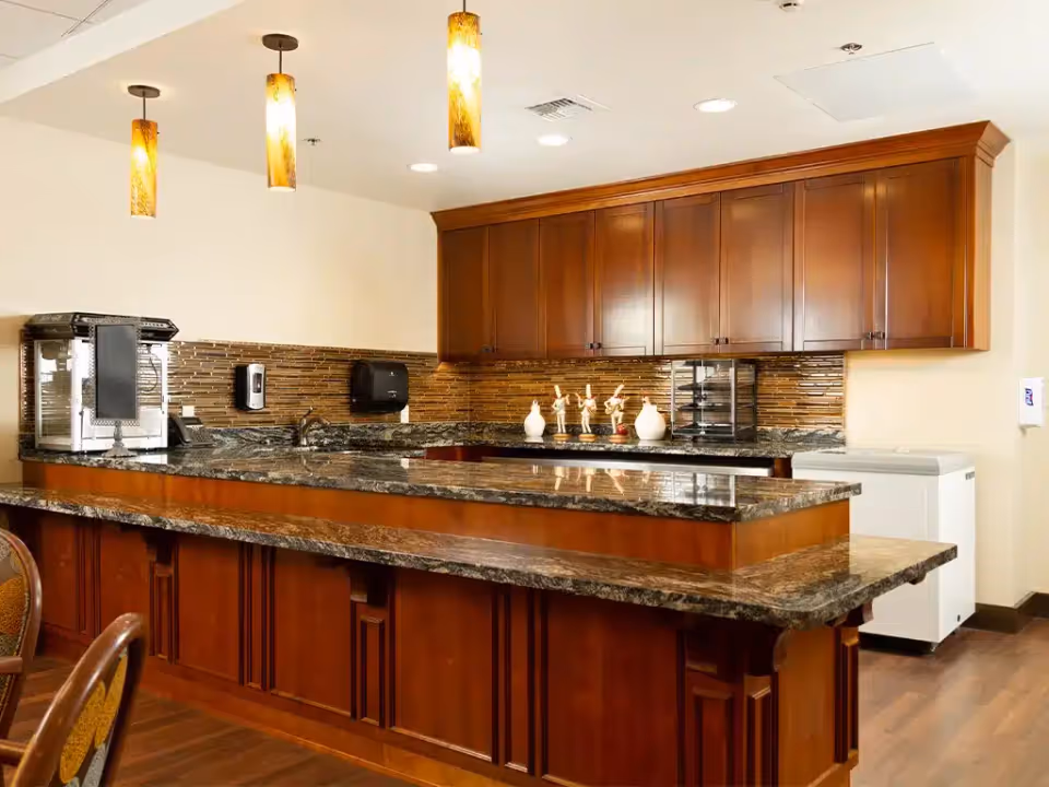 A communal kitchen serving counter with dark granite countertops, wooden cabinetry, pendant lights, and small appliances.