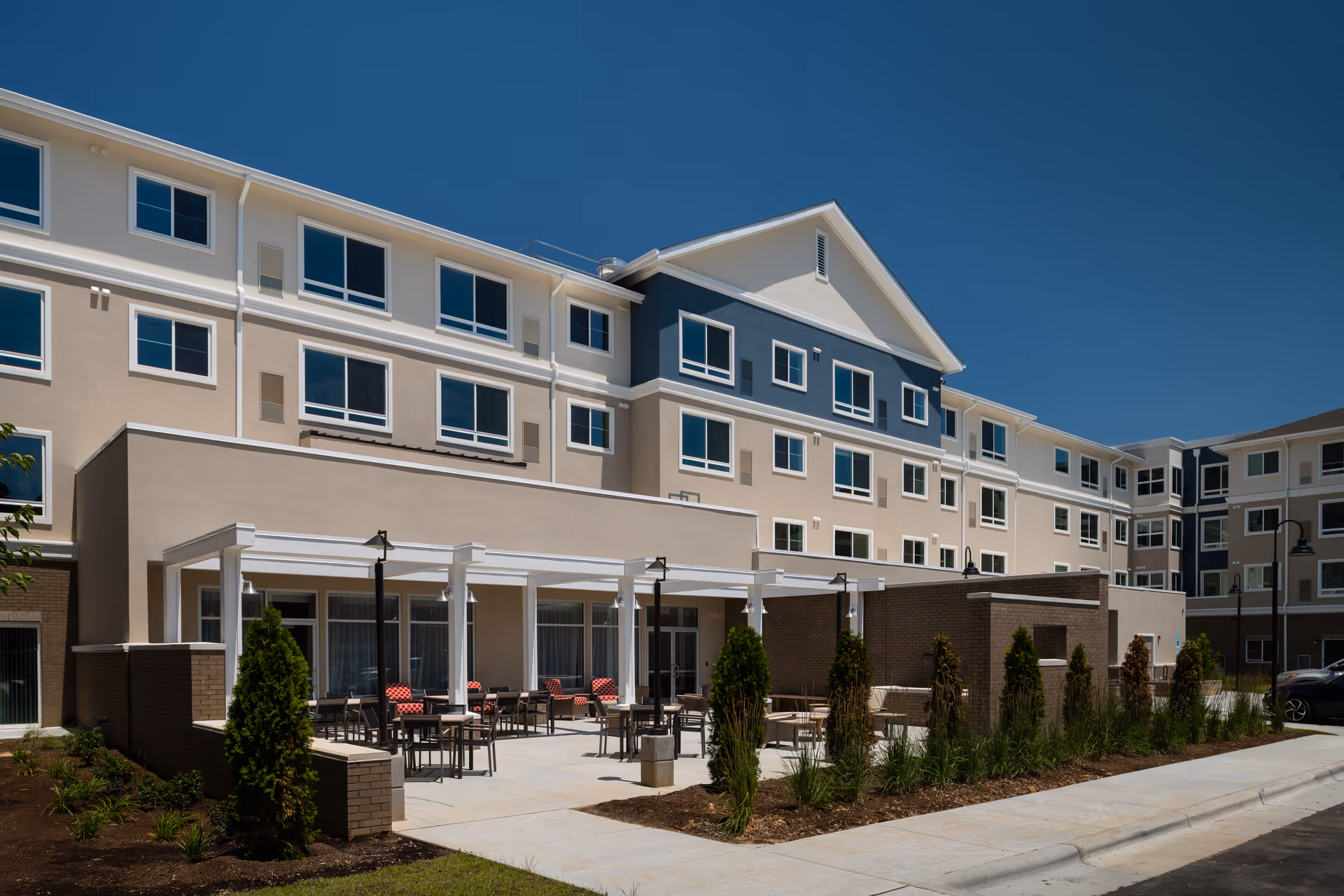 Exterior view of a multi-story senior living facility building with beige and blue walls, multiple windows, and a covered outdoor patio area with tables and chairs. The patio is surrounded by landscaped greenery and small trees under a clear blue sky.