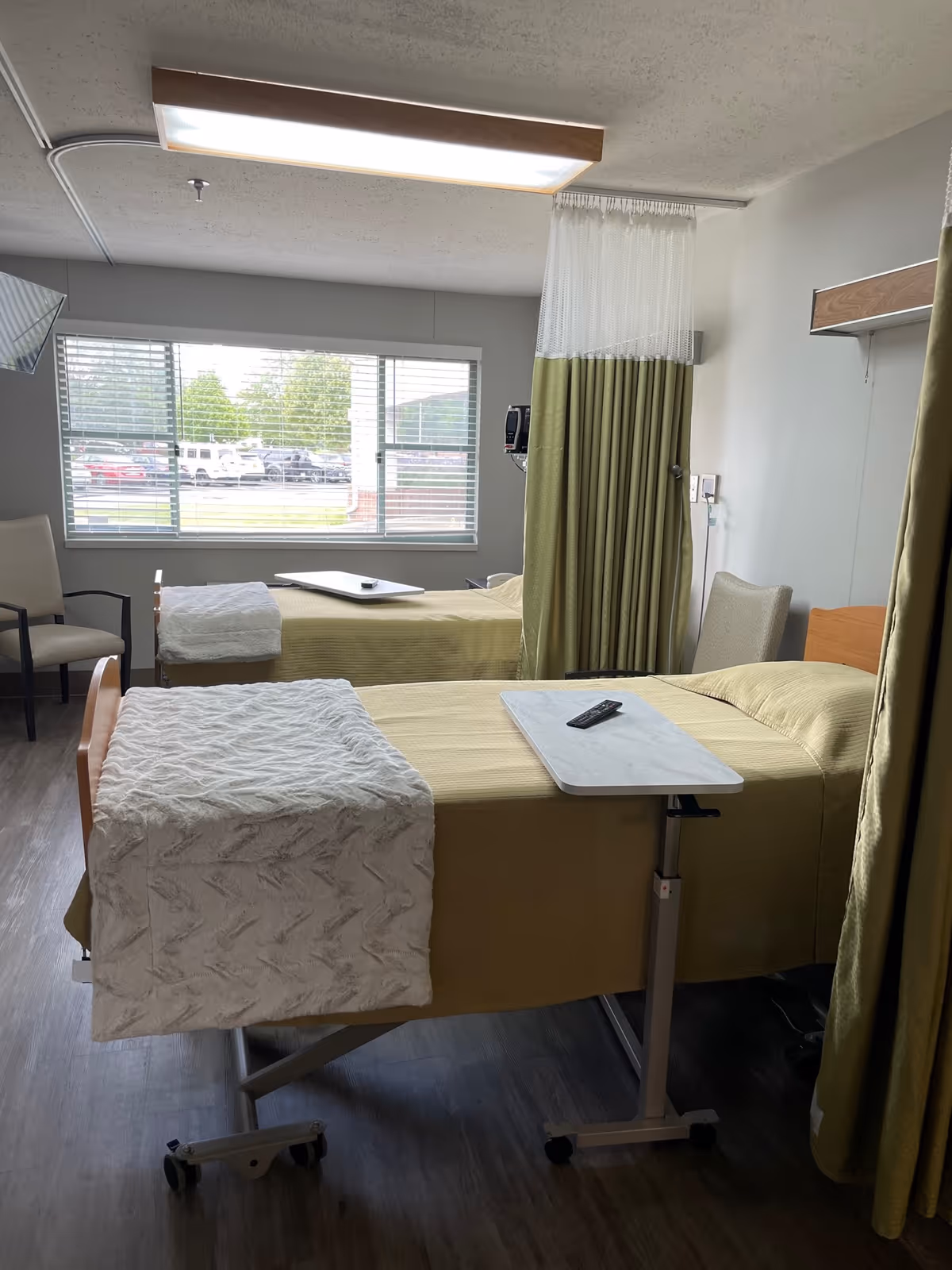 A room with two hospital-style beds, each covered with a yellow blanket and a white textured throw at the foot. Each bed has an overbed table with a remote control on it. There are two chairs near a large window with blinds, and a green privacy curtain partially drawn between the beds. The floor is wood-style, and the ceiling has a rectangular light fixture.