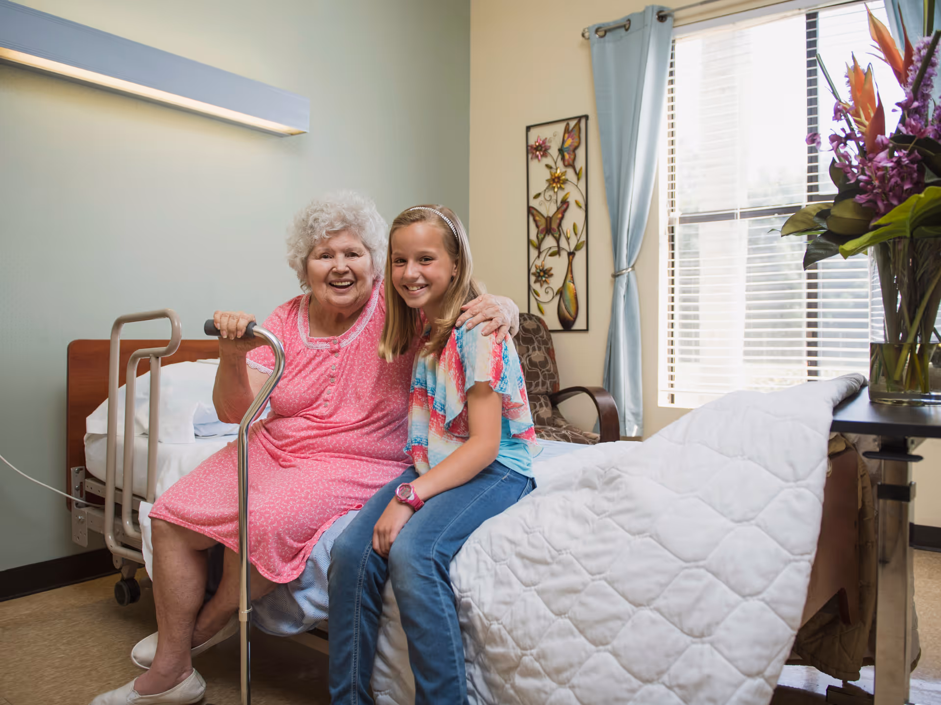 An elderly woman in a pink dress sitting on a hospital-style bed with a cane, smiling and hugging a young girl in a colorful shirt and jeans who is sitting beside her. The room has a window with blue curtains, a floral wall decoration, a chair, and a table with a vase of flowers.