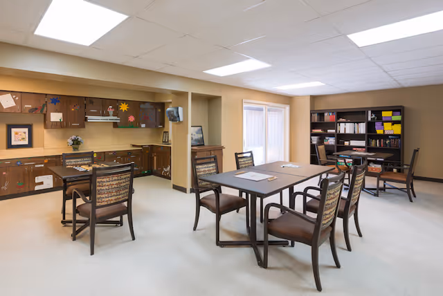 A bright communal activity room with multiple tables and chairs, cabinets decorated with crafts, and a bookshelf.
