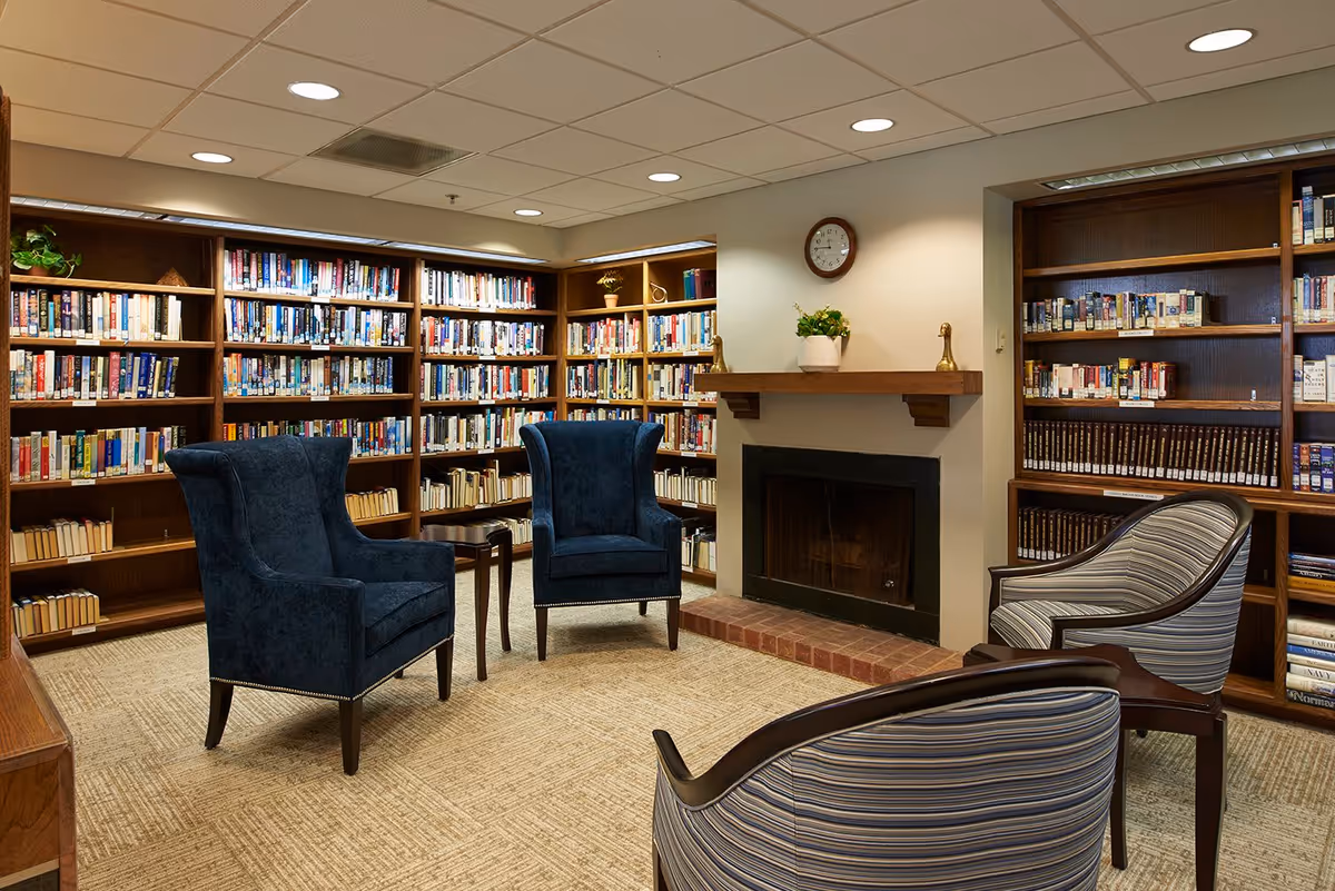 A cozy library room with wooden bookshelves filled with books lining the walls. There are four upholstered chairs arranged around a small wooden table in the center. A fireplace with a wooden mantle is on one wall, decorated with a potted plant and two small brass figurines. A clock hangs above the fireplace. The room has a carpeted floor and a ceiling with recessed lighting.