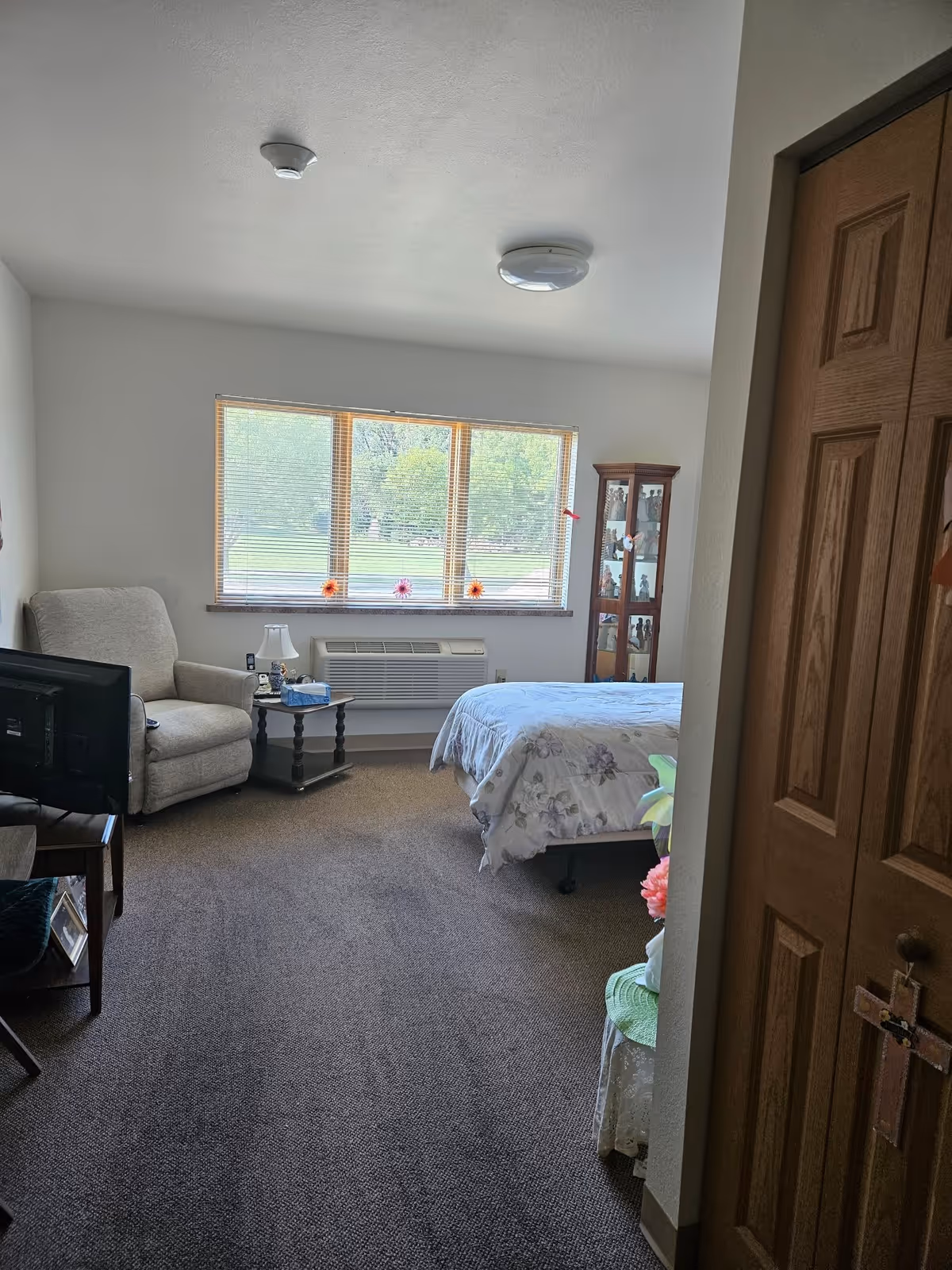 A cozy bedroom in Northview Assisted Living featuring a bed with floral bedding, a comfortable armchair next to a small side table with a lamp and tissues, a TV on a stand, a wooden cabinet with glass doors displaying figurines, and a large window with blinds overlooking a green outdoor area. The room has carpeted flooring and a wooden door with a cross hanging on it.