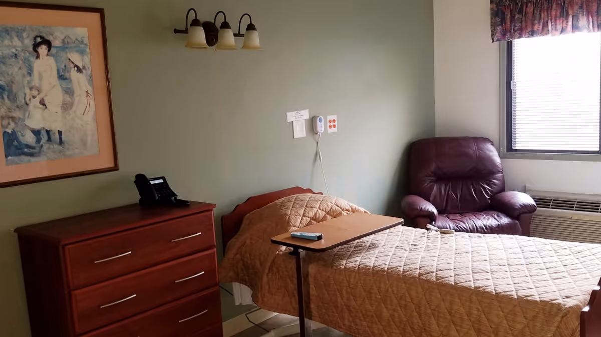 A small bedroom in a senior living facility with a single bed covered by a tan quilted blanket, a wooden dresser with a telephone on top, a maroon leather armchair, a window with blinds and floral valance, and a framed painting on the wall.