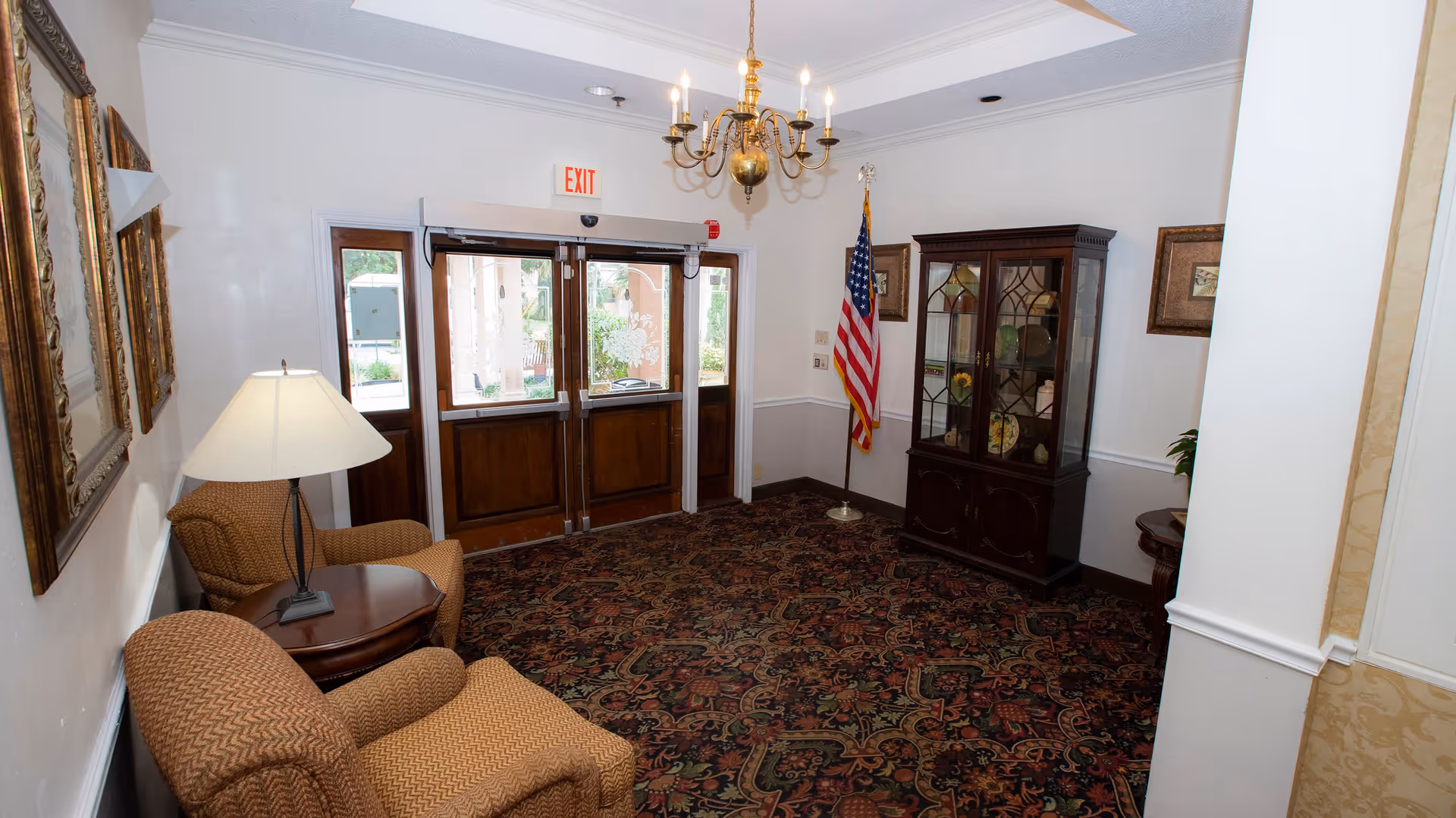 An interior view of a sitting area near an exit door in Colonial Assisted Living at Palm Beach. The space features two upholstered armchairs with a small wooden table and lamp between them, a patterned carpet, a chandelier hanging from the ceiling, framed artwork on the walls, an American flag in the corner, and a wooden display cabinet with glass doors containing decorative items.