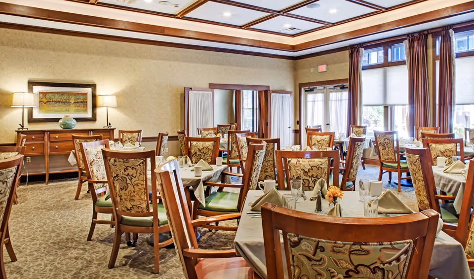 A well-lit dining room with multiple tables covered in beige tablecloths, each set with napkins, glasses, cups, and silverware. The chairs have wooden frames with patterned upholstery. The room features large windows with curtains, a wooden sideboard with lamps and a framed painting above it, and a coffered ceiling with recessed lighting.