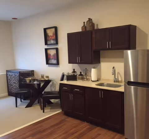 Interior view of a small kitchen area with dark wood cabinets, a stainless steel refrigerator, and a sink. Adjacent to the kitchen is a dining area with a round glass table, two chairs, and a cushioned bench with a zebra pattern. Two framed abstract paintings hang on the wall above the dining area.
