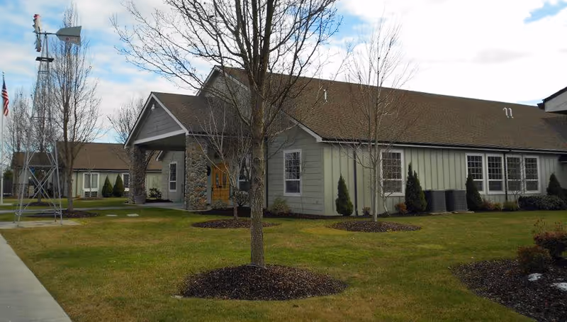 Exterior view of a single-story retirement lodge building with a covered entrance supported by stone pillars. The building is surrounded by a well-maintained lawn with young trees and shrubs. A metal windmill and an American flag are visible on the left side of the image under a partly cloudy sky.