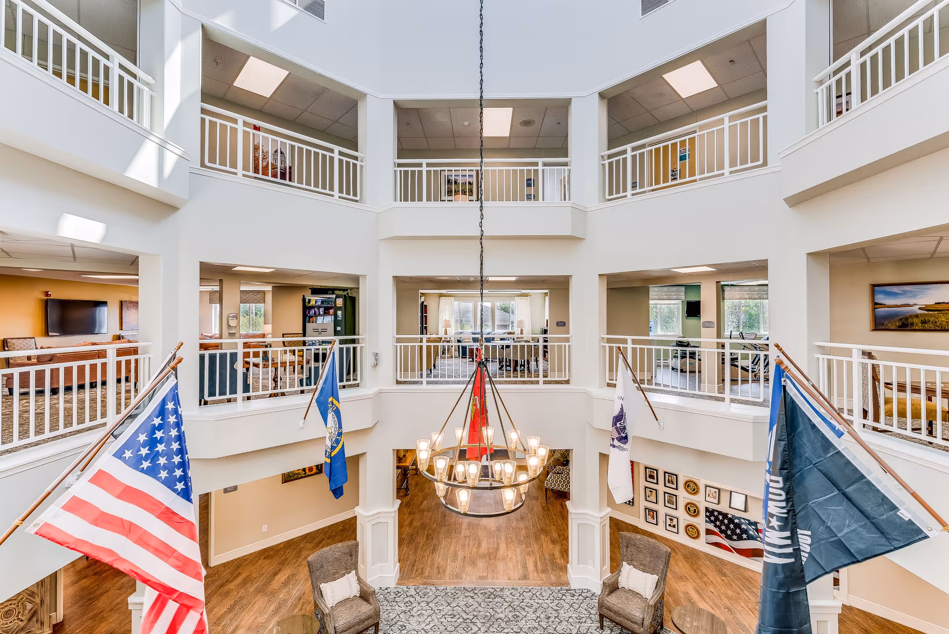 Interior view of a senior living facility's spacious common area with a high ceiling and balcony railings on the second floor. Four flags, including the American flag, are displayed prominently. The area features a large chandelier, seating with chairs and tables, and wood flooring. Multiple rooms and seating areas are visible on both floors.