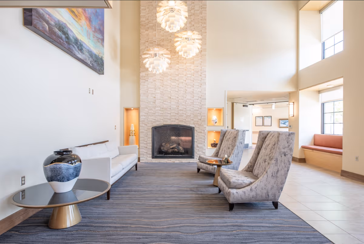 A bright and spacious living room area in a retirement community featuring a modern stone fireplace with three pendant lights above it. The room has a white sofa, two patterned armchairs, a round glass coffee table with a decorative vase, and a built-in bench seat by the window. The walls are light-colored, and there is a large colorful painting hanging above the sofa.