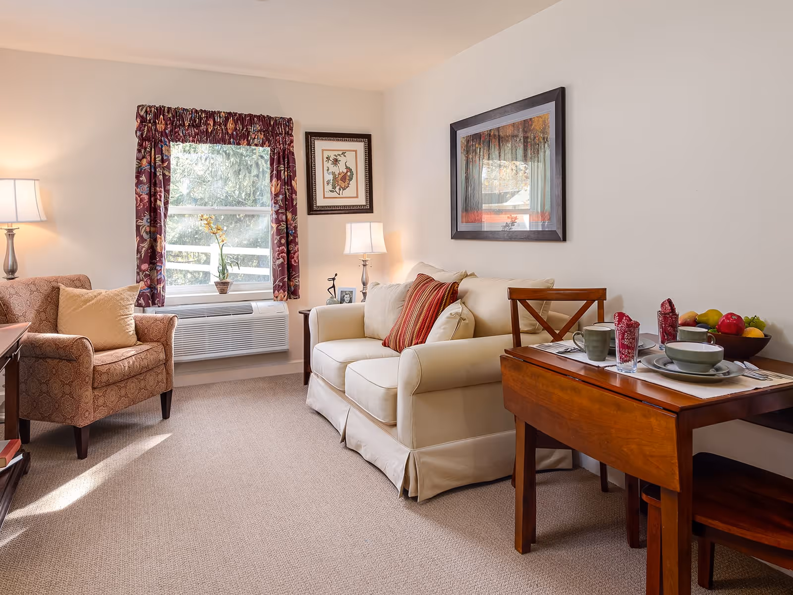 A cozy living room with a beige loveseat adorned with a red striped pillow, a patterned armchair with a beige cushion, and a wooden dining table set for two with dishes, cups, and red napkins. The room has beige carpeting, a window with floral curtains, two table lamps, and framed artwork on the walls.