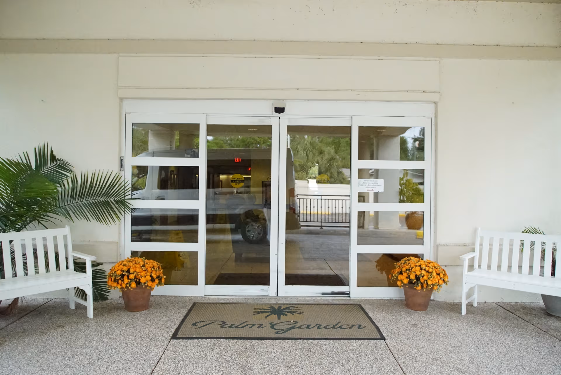 Front entrance of Palm Garden of Winter Haven with sliding glass doors, a welcome mat reading 'Palm Garden', white benches, and potted orange flowers.