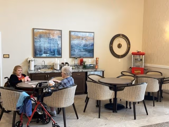 Two elderly women sitting at a round table in a common area with beige walls and tiled floor. One woman has a red walker beside her. Behind them is a counter with a sink, coffee maker, and other beverage supplies. On the wall above the counter are two abstract paintings and a large decorative clock. To the right, there is a popcorn machine and additional round tables with chairs.