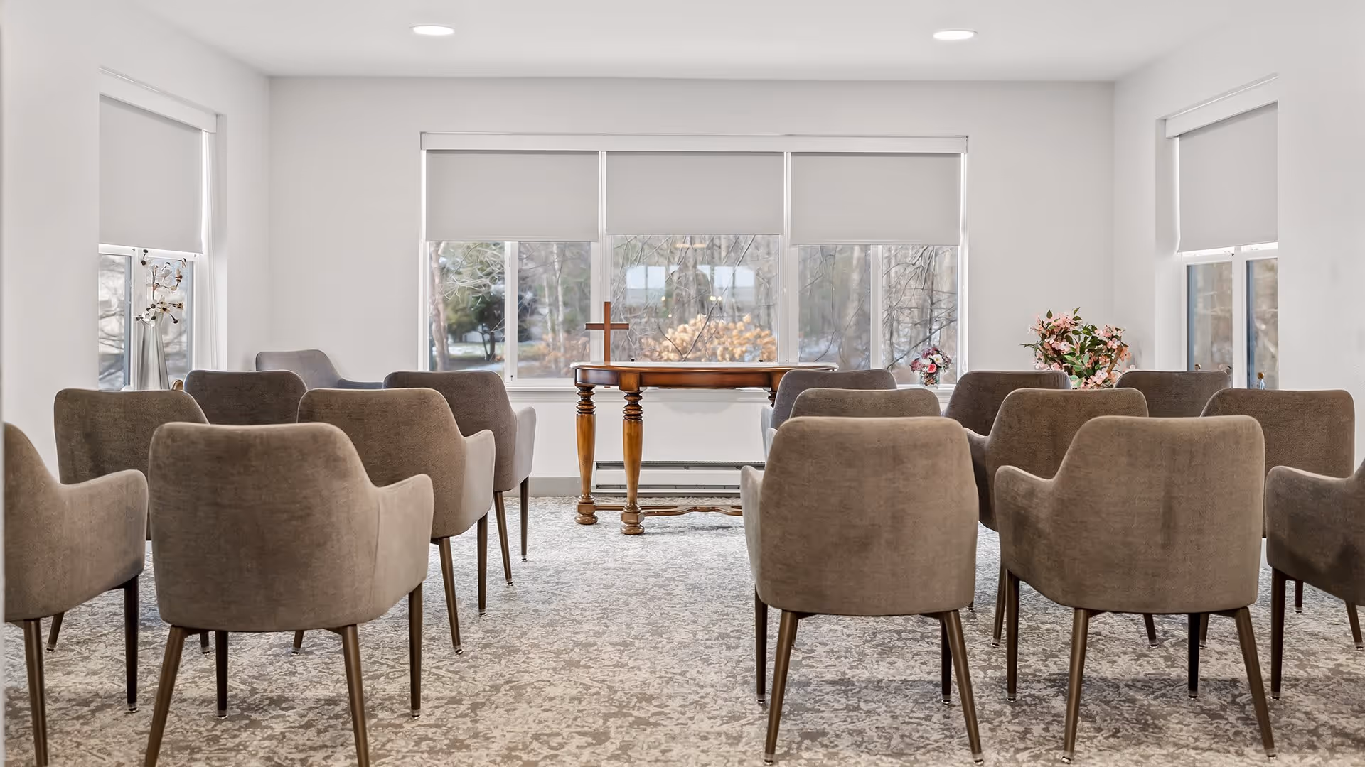 A small meeting or chapel room with rows of beige upholstered chairs facing a wooden table with a cross on it. The room has large windows with white roller blinds and light gray patterned carpet. There are flower arrangements on the table and on a side table near the windows.