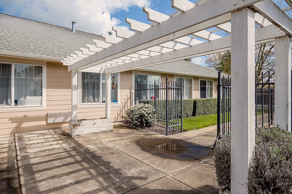 Courtyard with a white pergola casting shadows over a concrete patio, a black metal gate and walkway, and a beige single-story building with windows.