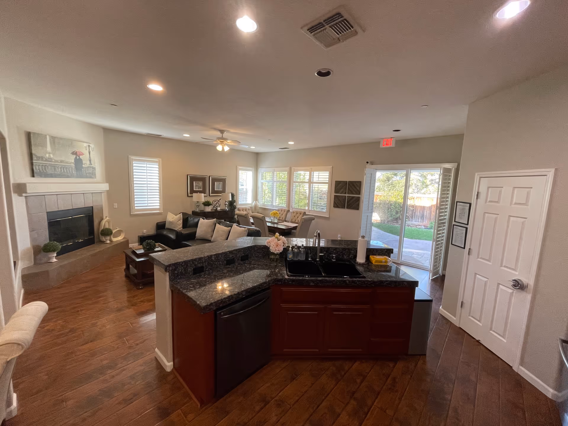 Interior view of a senior living facility showing an open living area with a kitchen island in the foreground, a dining table with chairs near windows, a seating area with couches and a fireplace, and a glass door leading to an outdoor space.