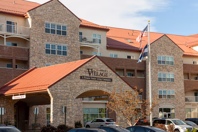 Exterior view of St. Andrew's Village senior living community building with stone and brick facade, red roof, multiple windows, balconies, and an entrance canopy. Two flagpoles with American and state flags are visible in front of the building, along with parked cars and a small tree.