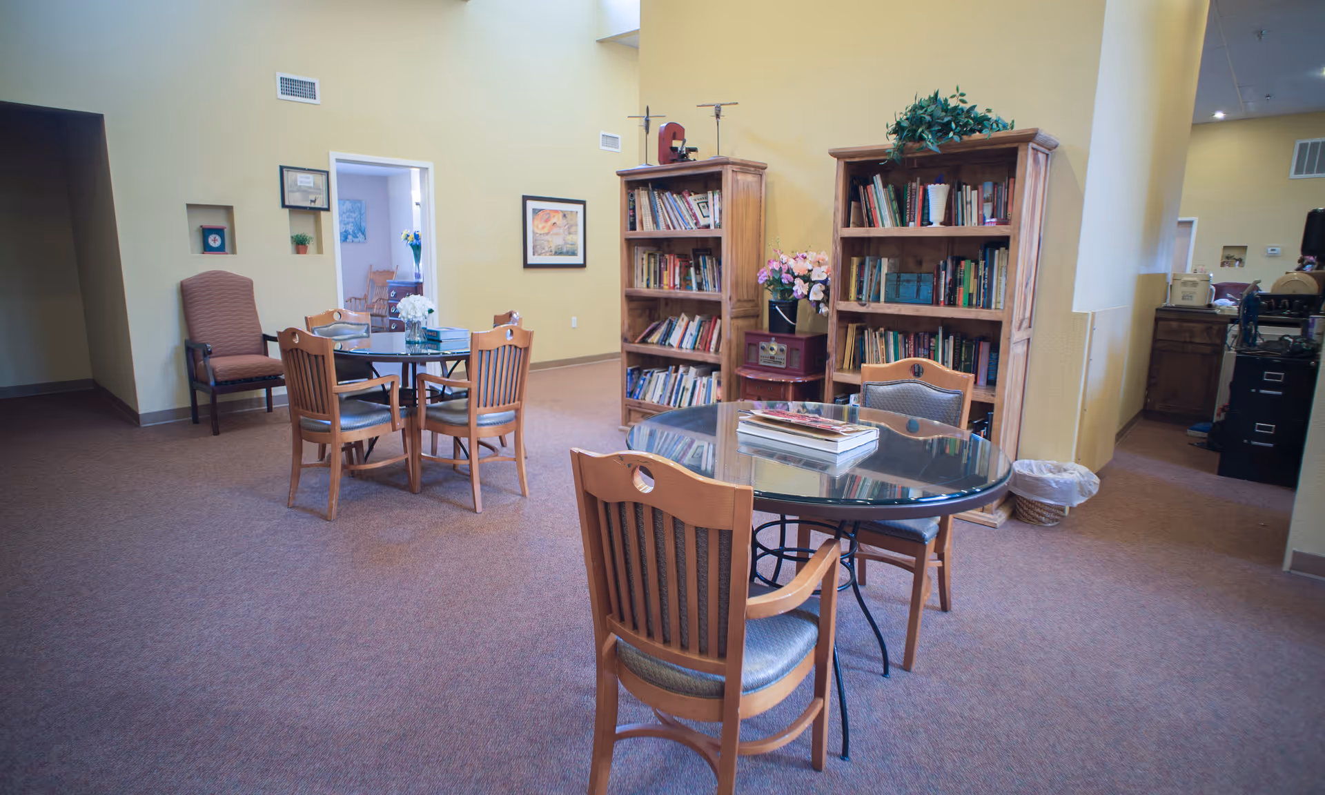 A cozy common area in a senior living facility with two round glass tables surrounded by wooden chairs. There are two wooden bookshelves filled with books and decorative items against a yellow wall. A comfortable armchair is placed near a doorway leading to another room with additional seating and decor. The carpet is brown, and the walls are painted light yellow.