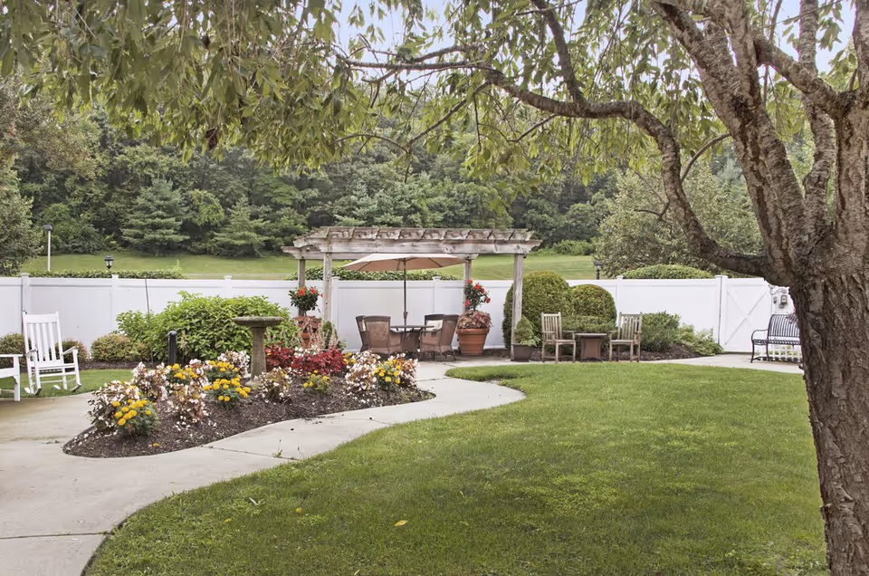 A peaceful outdoor garden area with a curved concrete pathway, flower beds with colorful flowers, a birdbath, and various seating options including white rocking chairs, wooden chairs, and a table with an umbrella under a wooden pergola. The area is surrounded by a white fence and lush green trees in the background.
