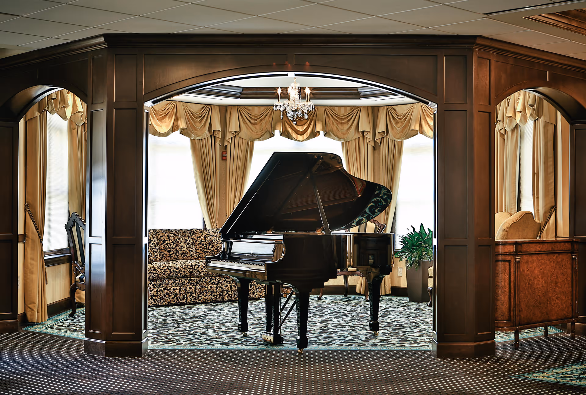 Elegantly furnished common room with a grand piano centered under an archway, surrounded by upholstered seating and drapery.