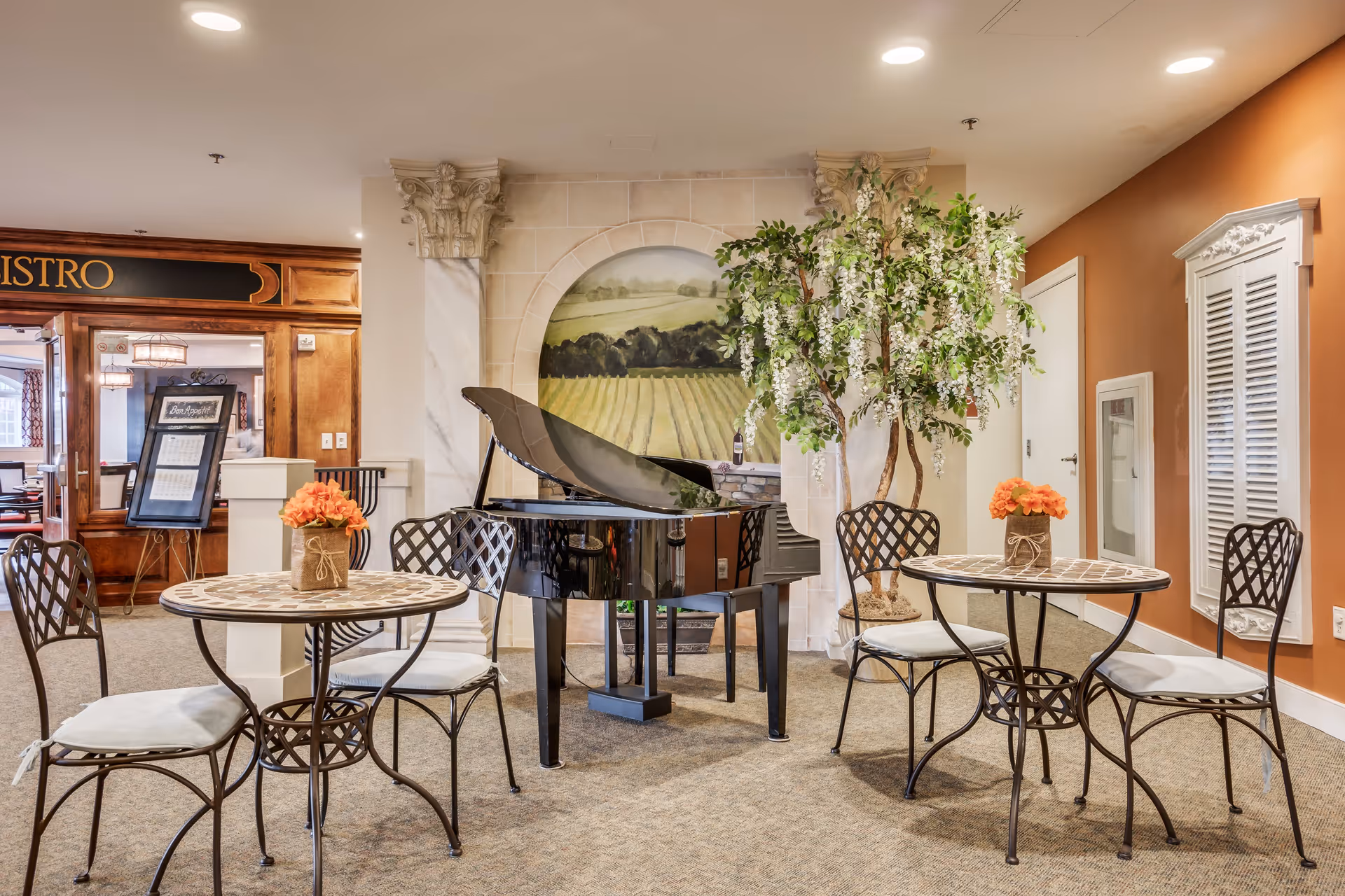 A cozy indoor seating area with two small round tables, each surrounded by two cushioned wrought iron chairs. A black grand piano is positioned in the center against a wall with a painted mural of a vineyard landscape. To the right, there is a tall artificial tree with white flowers. The walls are painted in warm tones, and the entrance to a bistro is visible in the background.