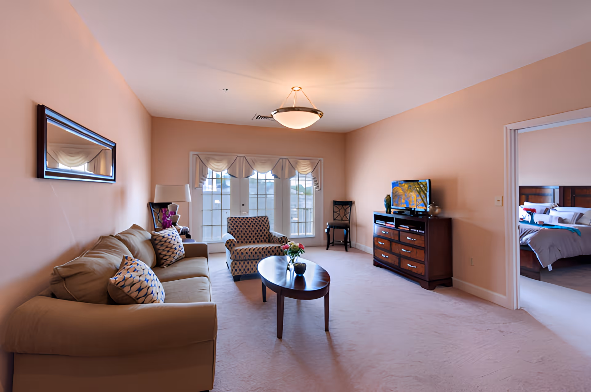 A cozy living room with peach-colored walls, a beige sofa with patterned pillows, a patterned armchair, a wooden coffee table with a small flower arrangement, a wooden dresser with a TV on top, and a window with decorative valances. A doorway leads to a bedroom with a neatly made bed.