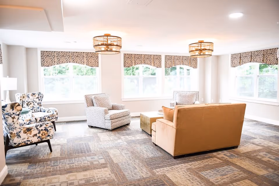 A bright and spacious living room area with large windows covered by patterned valances. The room features a beige sofa, two patterned armchairs, and two floral armchairs arranged around a central coffee table. The floor is carpeted with a geometric pattern, and two decorative ceiling lights hang from the ceiling.