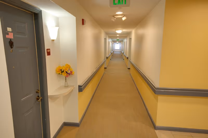 A long, well-lit hallway in a senior living facility with beige carpet and yellow and white walls. There are handrails along both sides of the hallway and a gray door on the left with a small American flag sticker and a room number plaque. A small shelf with a vase of yellow and purple flowers is mounted on the wall near the door. An exit sign is visible on the ceiling at the far end of the hallway.