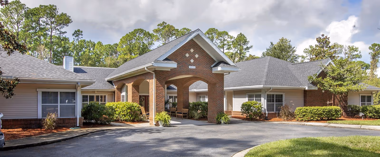 Exterior view of a single-story senior living facility building with a covered entrance supported by brick columns, surrounded by green bushes and trees under a partly cloudy sky.