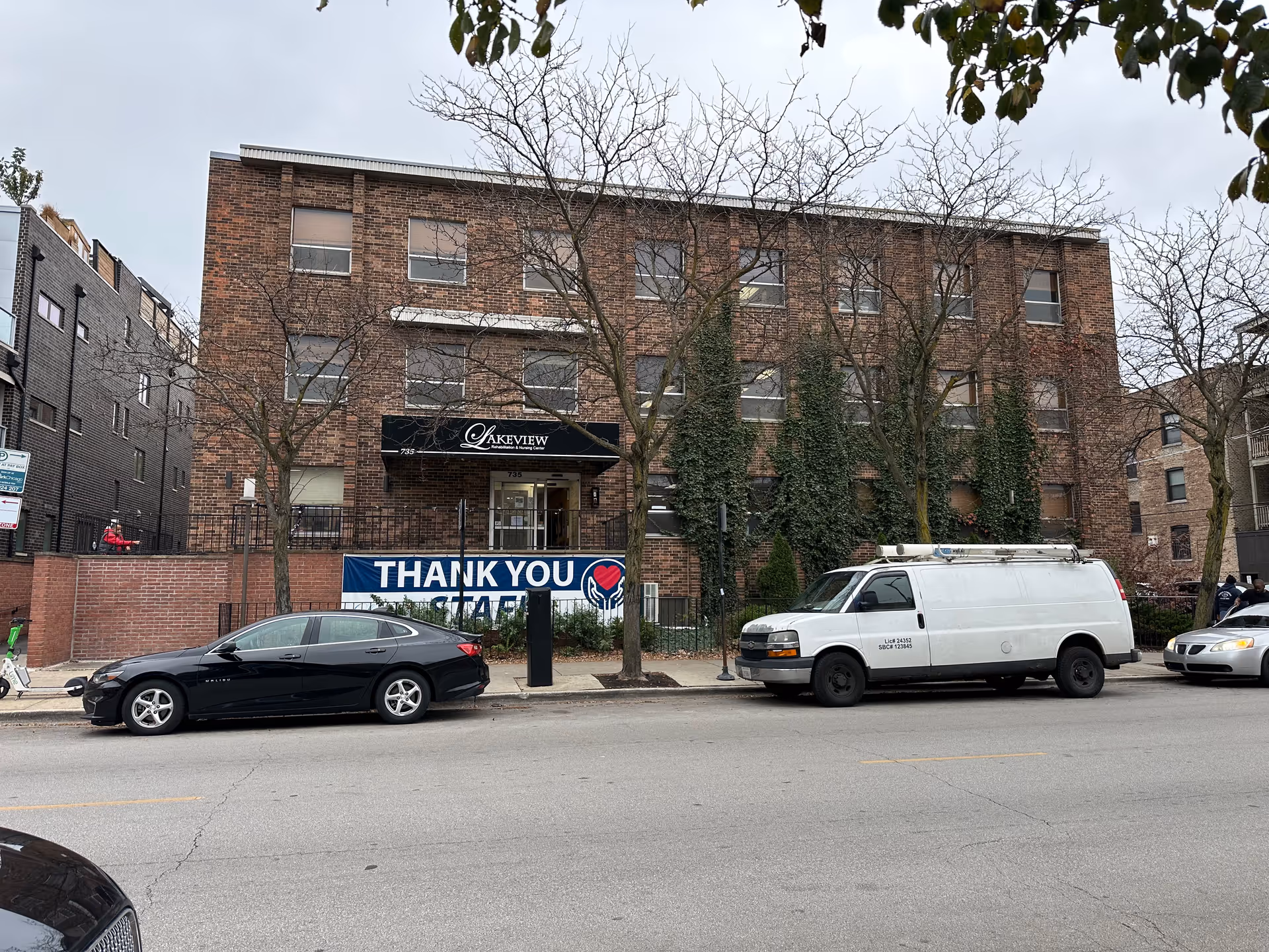 Exterior view of a three-story brick building with a black awning displaying the name 'Lakeview Rehabilitation & Nursing Center'. There are leafless trees and ivy growing on the building facade. Several cars are parked along the street in front of the building, and a large banner on the fence reads 'THANK YOU'.
