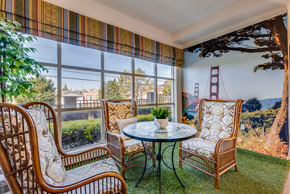 A cozy indoor seating area with three cushioned wicker chairs around a round glass-top table. The floor is covered with green artificial grass, and a large window reveals an outdoor view with trees and buildings. One wall features a large mural of the Golden Gate Bridge surrounded by trees and greenery.