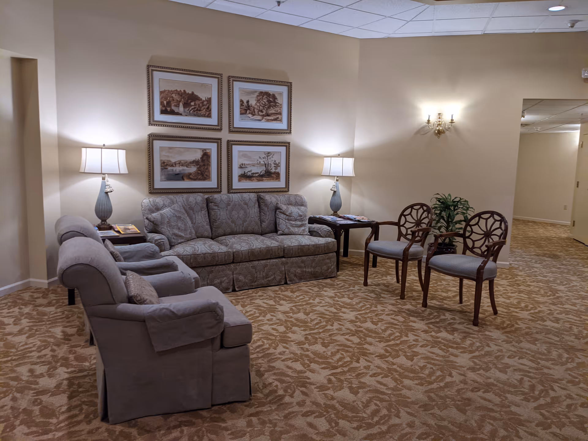 A cozy senior living community lounge area with a patterned sofa, two armchairs, two wooden chairs with cushioned seats, two side tables with lamps, framed landscape artwork on the wall, a potted plant, and beige patterned carpet.