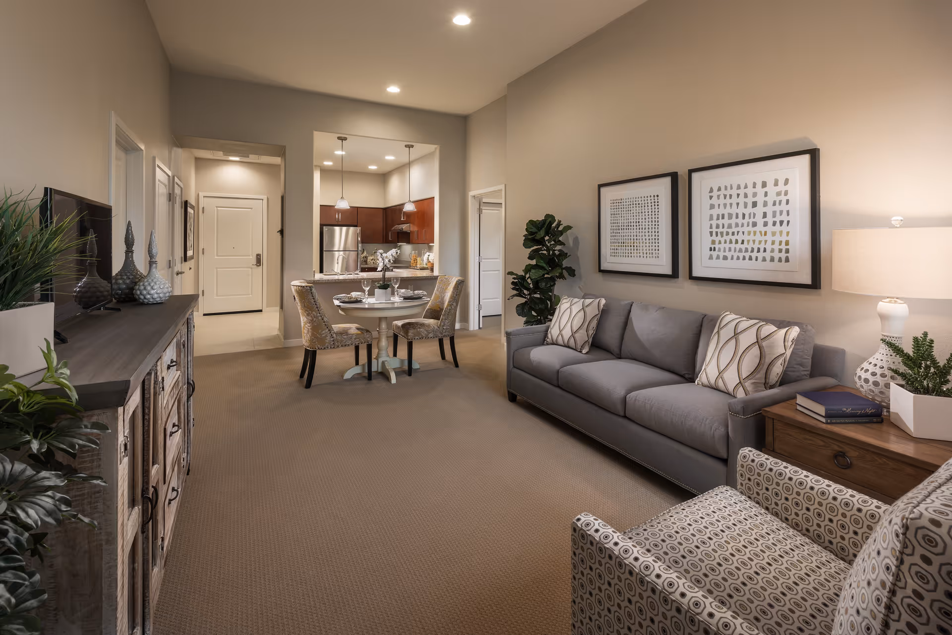 A cozy living room and dining area in a senior living facility. The living room features a gray sofa with patterned pillows, a patterned armchair, a wooden side table with a lamp and plants, and two framed abstract artworks on the wall. The dining area has a round table set for two with upholstered chairs. The kitchen with stainless steel appliances is visible in the background. The space is warmly lit with recessed ceiling lights and pendant lights over the kitchen counter.