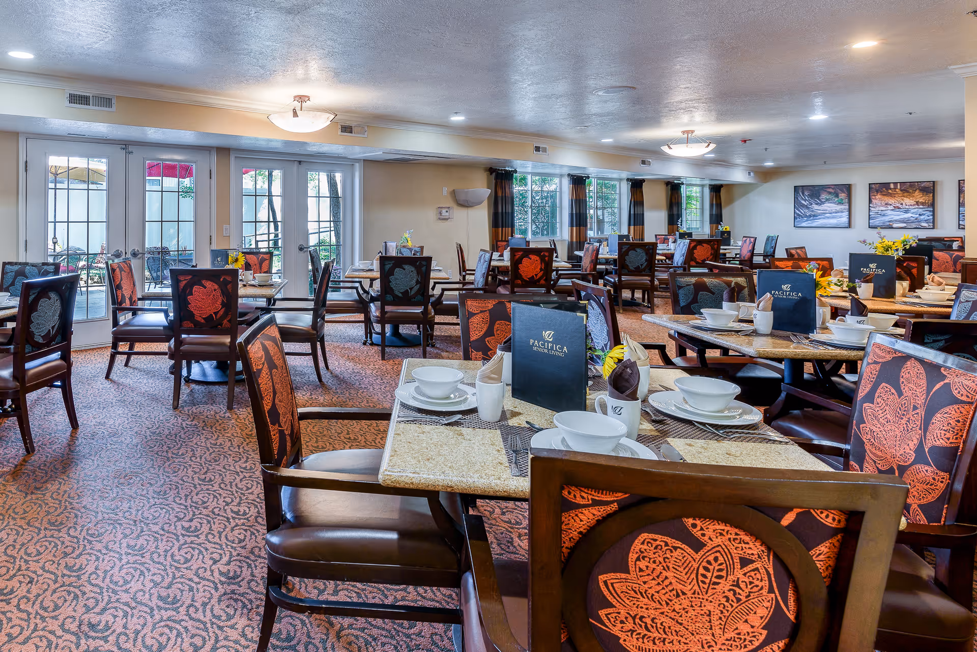A spacious dining room with multiple tables and chairs arranged neatly. Each table is set with white bowls, plates, cups, and napkins. The chairs have a floral patterned upholstery in shades of red and orange. Large windows and glass doors allow natural light to fill the room, and there are framed pictures on the walls. The room has a warm and inviting atmosphere.