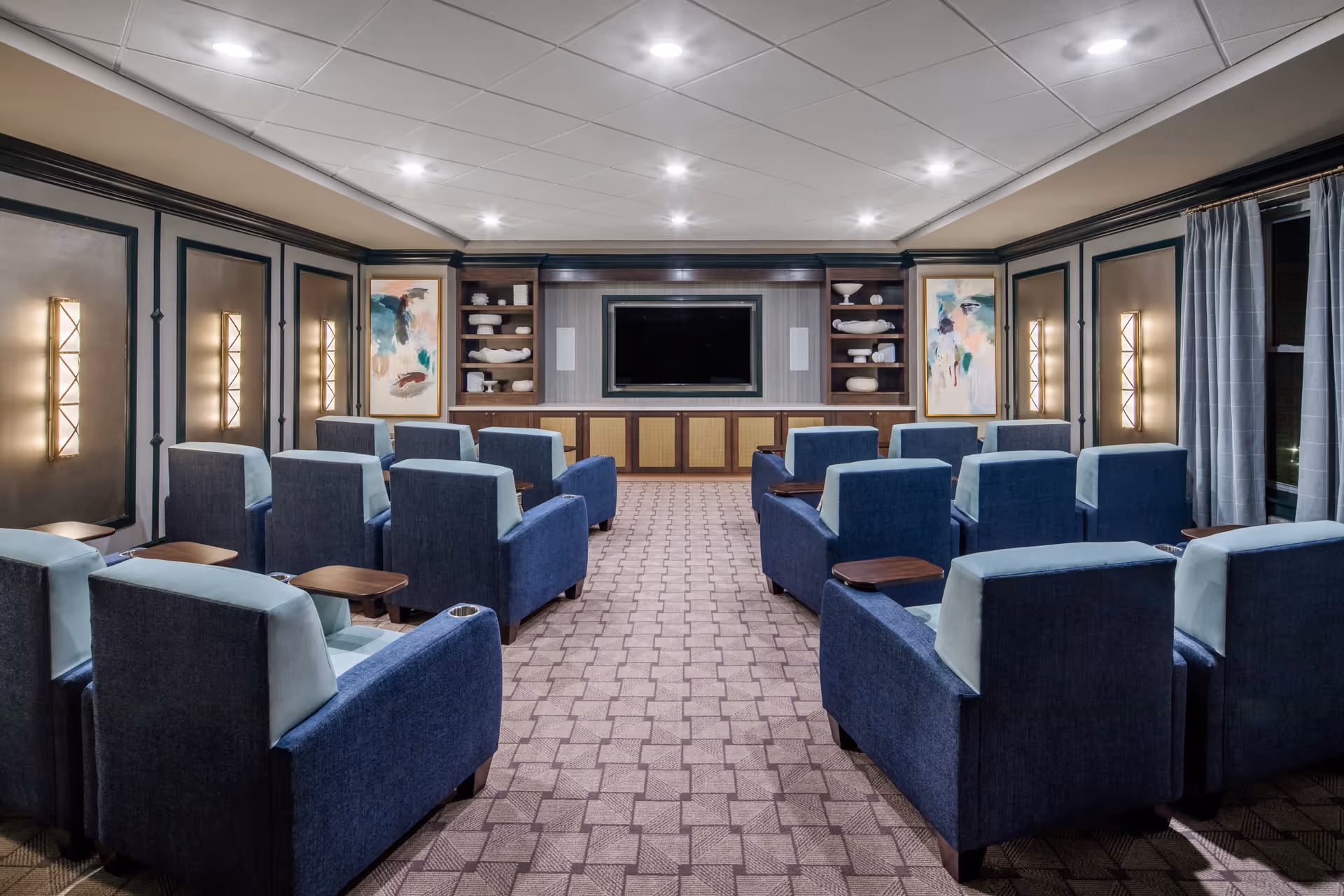 A cozy media room with multiple rows of blue and light gray armchairs facing a large flat-screen TV mounted on a wall with built-in shelves displaying decorative items. The room features soft lighting from wall sconces and recessed ceiling lights, with patterned carpet flooring and curtains on the windows.