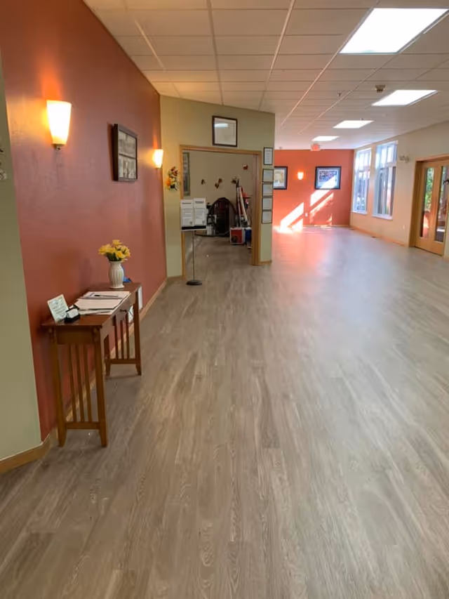 A spacious hallway with light wood flooring and a red accent wall on the left side. The wall has two sconces and a small wooden table with a vase of yellow flowers and some papers. The hallway leads to a room with cleaning equipment visible. Large windows on the right side let in natural light, and there are framed pictures on the walls.