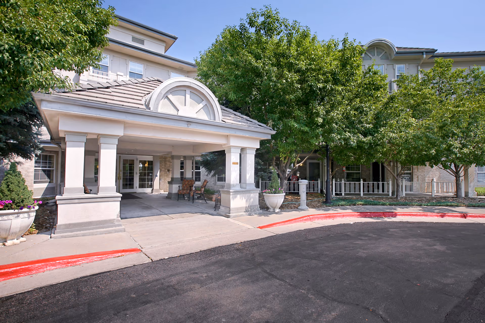 Exterior view of Belleview Suites at DTC showing the entrance with a covered driveway supported by columns, outdoor chairs, trees, and a paved driveway with a red curb.